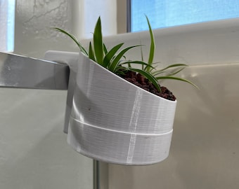 May include: White plastic plant hanger with a small green plant inside. The hanger is attached to a white surface.