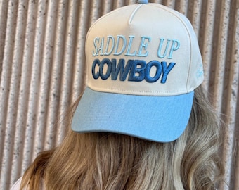 May include: A cream-colored baseball cap with a light blue brim. The cap features the embroidered text "SADDLE UP COWBOY" in blue. The hat is worn by a person with long blonde hair.