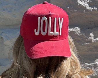 May include: A red baseball cap with the word "JOLLY" in white block letters. The cap is made of a textured fabric and has a curved brim. The cap is worn by a person with blonde hair.