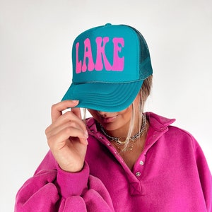 May include: Teal trucker hat with the word "LAKE" in pink, featuring a mesh back. The hat is worn with a pink long-sleeved shirt.
