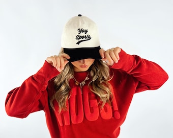 May include: A red sweatshirt with the word "HUGS!" in raised lettering. The person is wearing a white and black baseball cap with the words "Yay Sports" embroidered on the front. The person is also wearing a denim skirt.