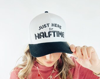 May include: White and black baseball cap with the text "JUST HERE for HALFTIME" in black. The cap is worn by a person wearing a red t-shirt with a graphic design and a gold necklace.