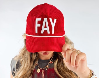 May include: A red corduroy baseball cap with the word "FAY" in white block letters. The hat has a white rope detail along the brim. The person is wearing a gray sleeveless shirt with a red graphic.