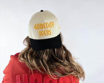 May include: A cream-colored baseball cap with the text "GAMEDAY BEERS" in gold lettering. The cap has a black brim and is worn by a person wearing a red sweatshirt with the word "HOGS!" in raised lettering.