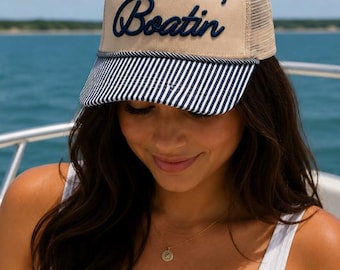 May include: Tan and navy blue trucker hat with the word "Boatin'" embroidered in navy blue script. The hat features a striped brim and a mesh back. A woman is wearing the hat on a boat.