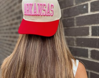 May include: A cream-colored baseball cap with a red brim and a red button on top. The word "ARKANSAS" is embroidered in pink across the front of the cap. The cap is worn by a person with long, light brown hair.