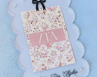 May include: A clear, scalloped-edge acrylic sign with a floral print card featuring the letters "ZTA" in a pink rectangle. A black bow is attached at the top. The words "Zeta Tau Alpha" are written in script at the bottom.