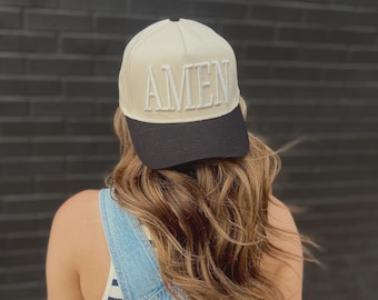 May include: A beige baseball cap with a black brim. The word "AMEN" is embroidered in white on the front of the cap. The cap is worn by a person wearing a striped tank top and denim overalls.