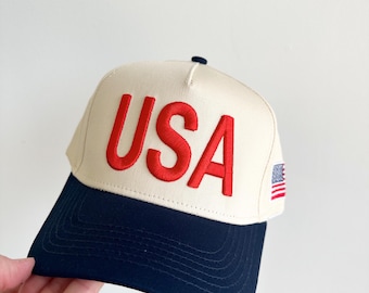 May include: Cream-colored baseball cap with a navy blue brim. The word "USA" is embroidered in red across the front. A small American flag patch is on the side. A classic, patriotic accessory.