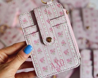 May include: Pink floral print cardholder with a gold key ring. The cardholder features a vertical stripe pattern with small pink roses and the Greek letters AXΩ. It has a snap closure and a zippered pocket.