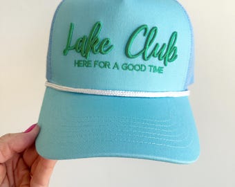 May include: A light blue trucker hat with a curved brim and mesh back. The hat features the embroidered text "Lake Club" in green script, with "HERE FOR A GOOD TIME" below. A white rope detail accents the brim.