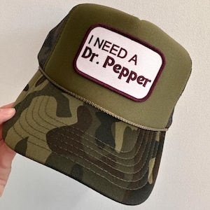 May include: A camouflage trucker hat with a green crown and a curved brim. The hat features a white patch with the text "I NEED A Dr. Pepper" in maroon lettering. The hat has a mesh back.