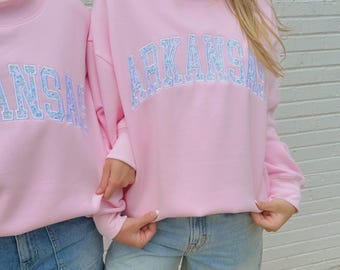 May include: Two light pink sweatshirts with the word "ARKANSAS" in light blue floral lettering. The sweatshirts have a mock neck and are paired with denim shorts. The background is a white brick wall.