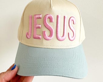 May include: A baseball cap with a light beige crown and a light blue brim. The word "JESUS" is embroidered in pink across the front. The cap has a button on top.