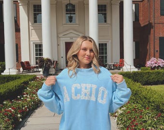 May include: A light blue sweatshirt with the text "A CHI O" in white floral lettering. The sweatshirt is being held up in front of a large white building with columns and a red brick building.
