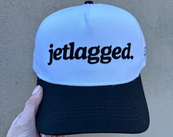 May include: White and black baseball cap with the text "jetlagged" embroidered in black on the front.