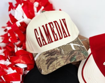 May include: A stack of baseball caps with the word "GAMEDAY" embroidered on the front in red or white. One cap has a camouflage brim. Red and white pom-poms are in the background.