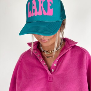 May include: Teal trucker hat with the word "LAKE" in pink, retro-style lettering. The hat has a mesh back and a curved brim. The person in the image is wearing a pink collared shirt.
