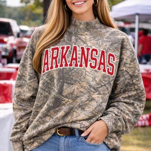 May include: A camouflage-patterned sweatshirt with the word "ARKANSAS" in red block letters. The sweatshirt has a mock neck and long sleeves. The person is wearing blue denim shorts and a brown belt with a gold buckle.