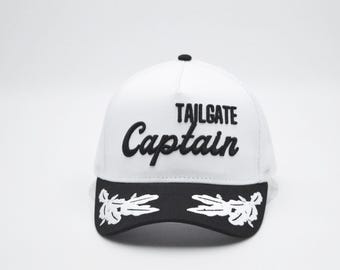 May include: White baseball cap with the words "TAILGATE Captain" embroidered in black. The brim is black with a white floral design. The cap has a black button on top.