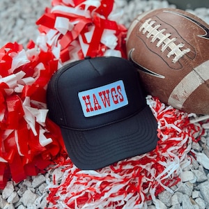 Puede incluir: Gorra de camionero negra con un parche azul y rojo que dice "HAWGS". La gorra está rodeada de pompones rojos y blancos y un balón de fútbol marrón.
