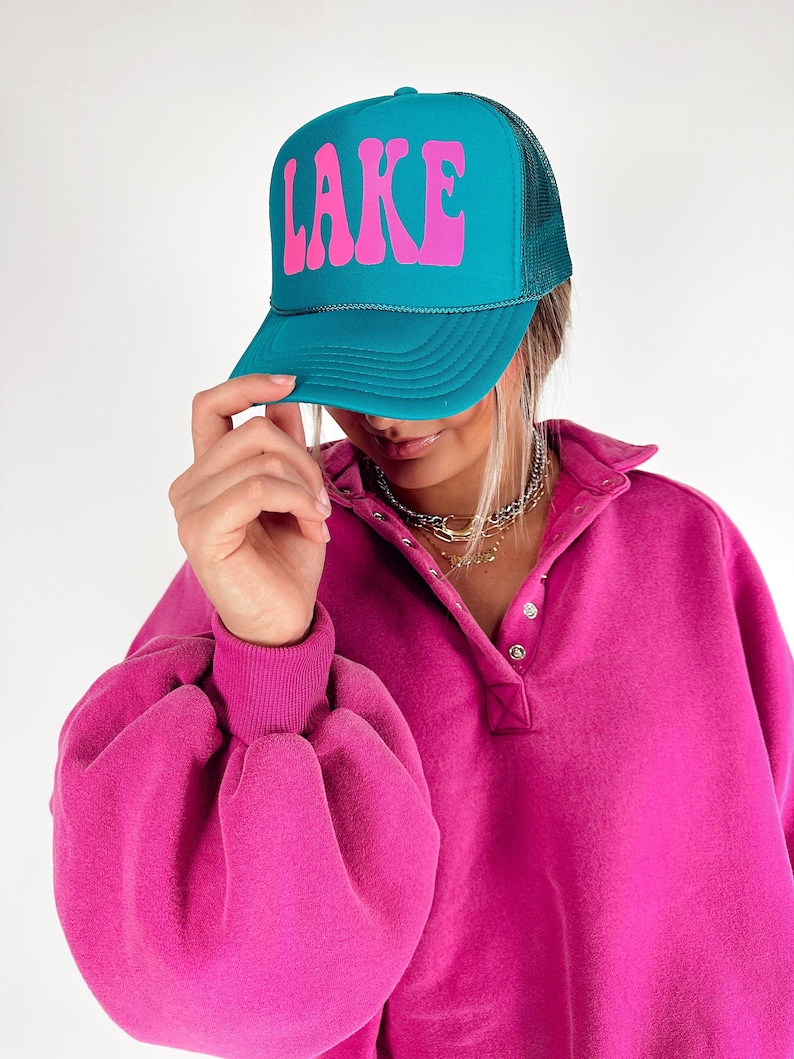 May include: Teal trucker hat with the word "LAKE" in pink, featuring a mesh back. The hat is worn with a pink long-sleeved shirt.
