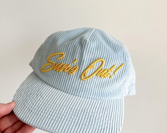 May include: Light blue corduroy baseball cap with the embroidered text "Sun's Out!" in gold. The hat has a curved brim and a structured crown. The corduroy fabric has vertical ridges.