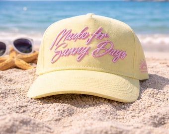 May include: A light yellow baseball cap with the embroidered text "Made for Sunny Days" in pink script. The cap is on a sandy beach with sunglasses and a starfish visible in the background. The ocean and sky are visible.