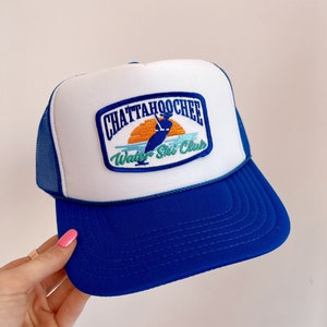 May include: A white and blue trucker hat with a patch that says "Chattahoochee Water Ski Club". The patch features a sunset, a skier, and a blue wave.