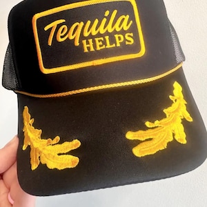 May include: Black trucker hat with a yellow embroidered patch that says "Tequila Helps". The hat also has two yellow embroidered designs on the sides.
