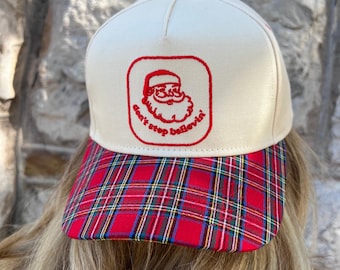 May include: A cream-colored baseball cap with a red plaid brim. The cap features a red square with a Santa Claus graphic and the text "don't stop believin'" in red cursive.