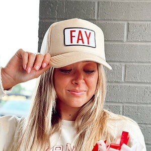 Puede incluir: Una gorra de camionero beige con un parche rojo que dice "FAY".