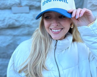 May include: A woman wearing a white puffer jacket and a beige and blue baseball cap with the text "tired mom."