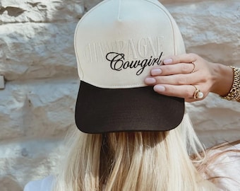 May include: A cream-colored baseball cap with a black brim. The cap features the embroidered text "CHAMPAGNE Cowgirl" in a script font. The cap is being held by a person with blonde hair, wearing rings and a gold watch.