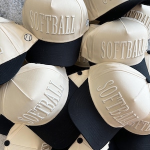 May include: A pile of cream-colored softball caps with black brims. The word "SOFTBALL" is embroidered in white on the front of each cap. Some caps have a small baseball graphic embroidered on the side.