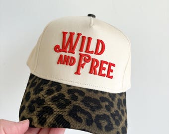 May include: A beige baseball cap with the embroidered words "WILD AND FREE" in red. The brim features a leopard print pattern in shades of brown and black. The cap is held by a hand.