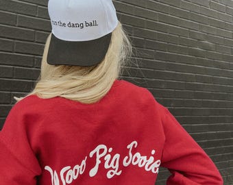 May include: Red sweatshirt with white cursive text that reads "Woo Pig Sooie" on the back. A person is wearing the sweatshirt and a white baseball cap with black brim that says "run the dang ball."