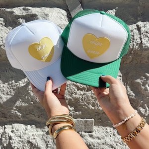 May include: Two white trucker hats with green mesh backs. Both hats feature a yellow heart with the text "chips & queso" in white.
