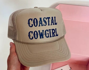 May include: A beige trucker hat with a blue embroidered design that says "COASTAL COWGIRL".
