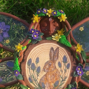 May include: A handmade woodland fairy figurine with a brown face, closed eyes, and a flower crown. The fairy is holding a wooden slice with a painted image of a brown hare in a field of blue thistles.