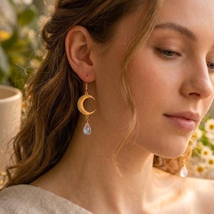 May include: A gold-toned earring featuring a crescent moon charm suspended from a hook, with a small, translucent, teardrop-shaped gemstone dangling from the bottom of the moon. The earring has a polished finish and a delicate, lightweight design.