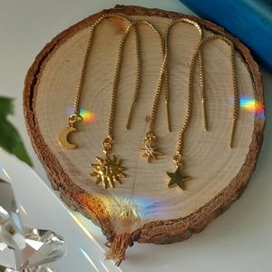 May include: Gold threader earrings with celestial charms. The earrings feature a crescent moon, a sun, a starburst, and a five-pointed star. The earrings are displayed on a wooden slice.