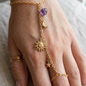 Puede incluir: Pulsera de cadena de mano dorada con un dije en forma de corazón púrpura, una luna creciente, un sol con una cara y una estrella. La pulsera se conecta a un anillo. También son visibles varios anillos dorados.