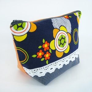 Könnte beinhalten: Eine marineblaue Reißverschlusstasche mit einem Retro-Blumenmuster in Gelb, Orange und Grün. Sie hat eine weiße Spitzenborte und orangefarbene Seitenteile. Der Reißverschluss ist dunkelgrau.