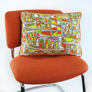 Peut inclure: Un fauteuil orange avec un coussin à motifs géométriques. Le coussin a un fond blanc avec des formes géométriques jaunes, orange et brunes.