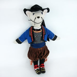 May include: A soft toy mouse dressed in traditional attire. The mouse features a black hat, blue jacket with red and gold detailing, a grey waistcoat, brown shorts, and black socks with red and white accents. The doll is set against a white backdrop.