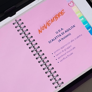 Könnte beinhalten: Eine digitale Planerseite auf einem Tablet-Bildschirm mit rosa Hintergrund und Spiralbindung. Der Text lautet NOVEMBRE, U.D.A. SCALETTA DI AGILITA' (4 lezioni), primo approccio alla scaletta, lavori di gruppo, workout musicale und verifica finale.