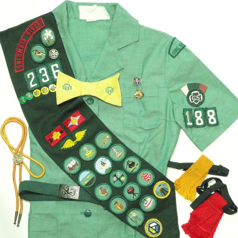 Puede incluir: Un uniforme verde de Girl Scout con una banda que presenta una variedad de insignias y parches. La banda tiene el texto "Columbia River" y el n&uacute;mero "236". El uniforme tambi&eacute;n tiene una corbata de lazo amarilla y una insignia con el texto "Girl Scouts" y el n&uacute;mero "188".