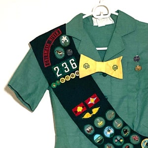 Puede incluir: Un uniforme verde de Girl Scout con una banda que presenta una variedad de insignias y una corbata de lazo. La banda tiene el texto "Columbia River" y el n&uacute;mero "236" en ella.