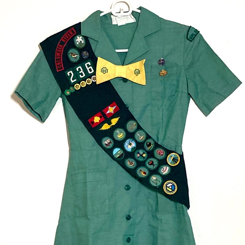 Puede incluir: Un vestido verde de uniforme de Girl Scout con mangas cortas y una banda con varias insignias y el texto "Columbia River 236".